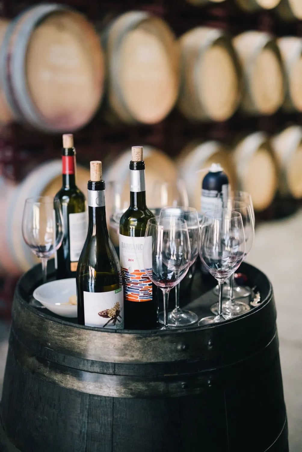 Wine bottles and glasses on a wooden barrel in a winery, with a plate of snacks in the foreground.