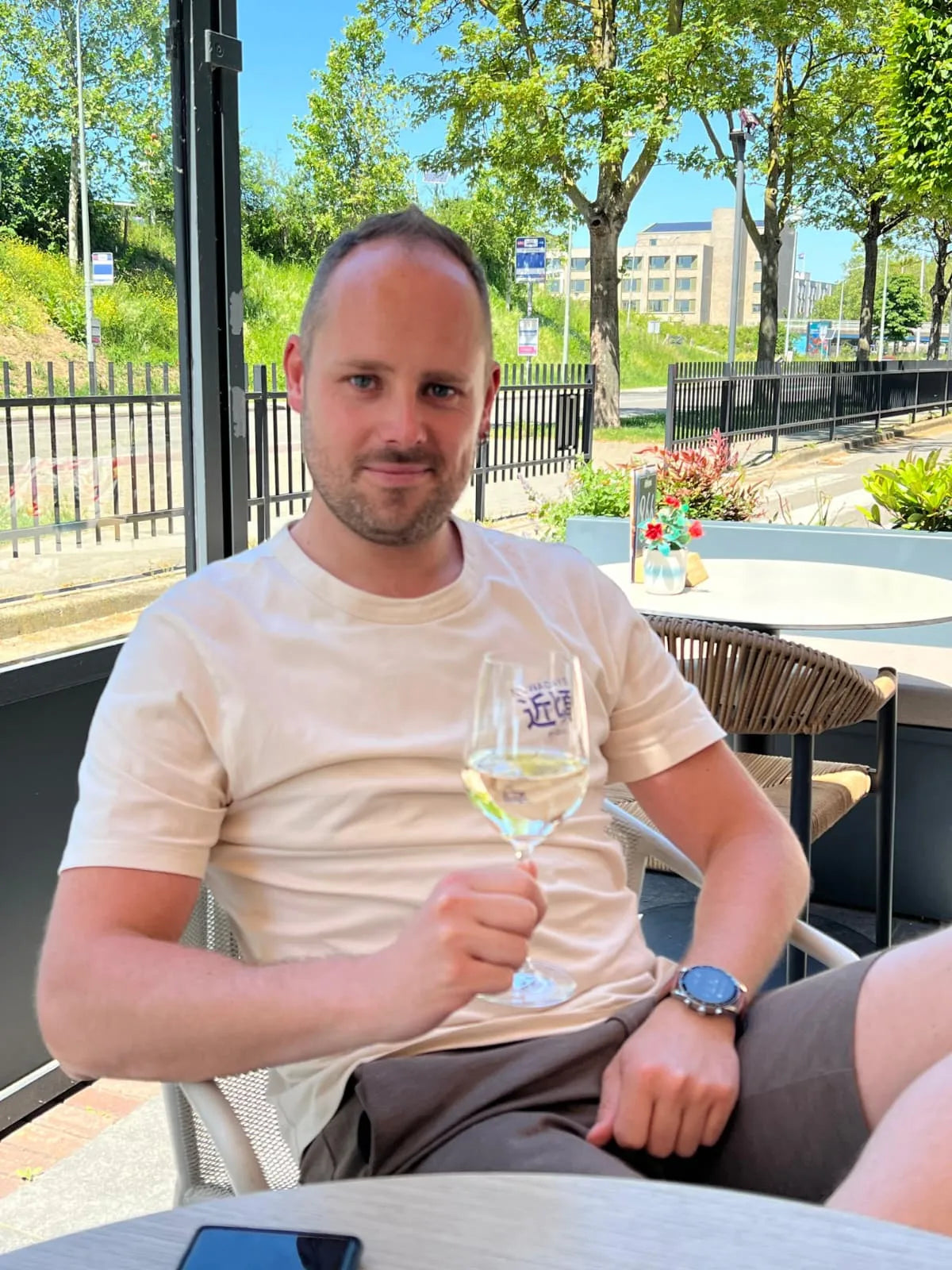 A man sitting at a table outdoors, holding a glass of white wine, wearing a casual t-shirt and shorts.